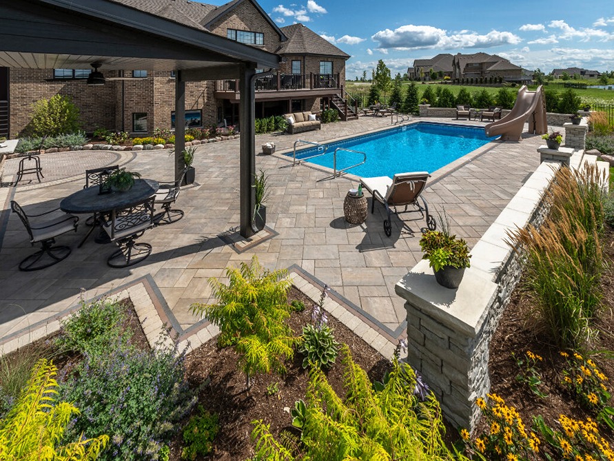 Wide angle view of a backyard pool with a paver deck, pavilion, stone walls and softscaping.